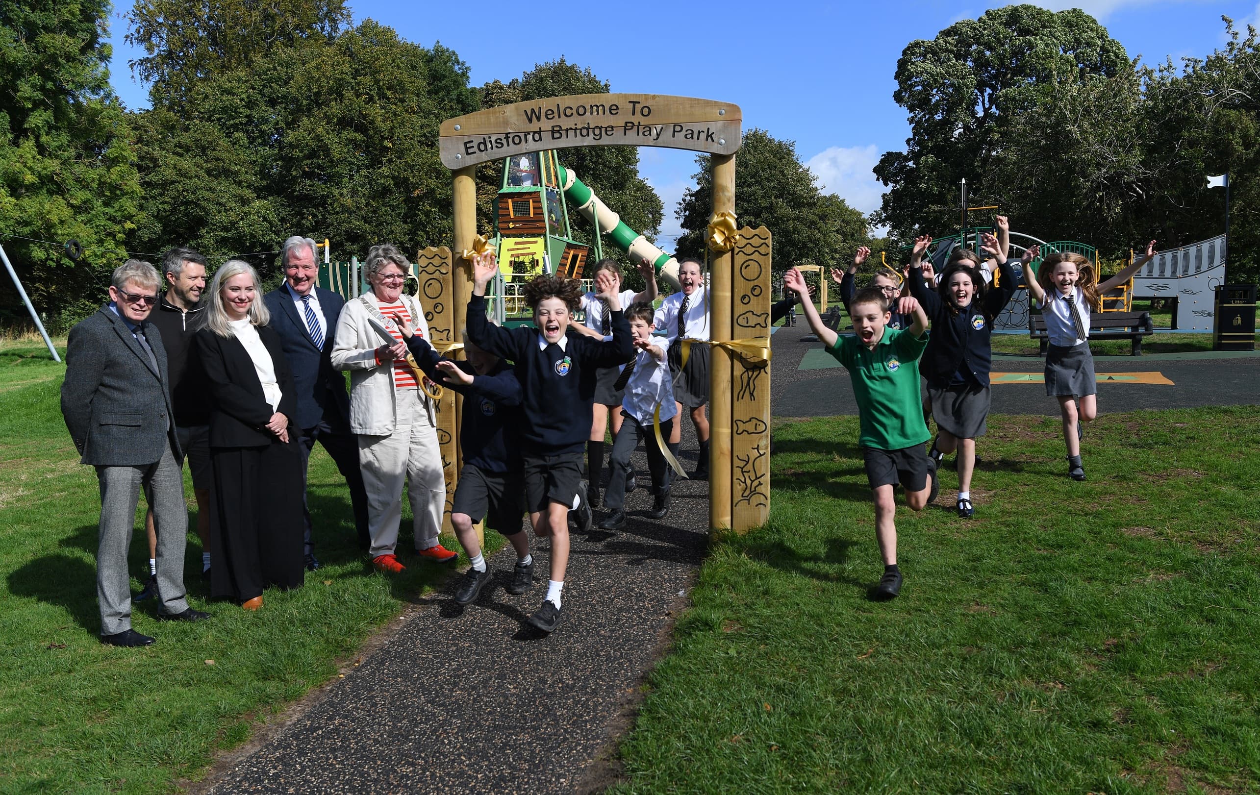 Young children and Ribble Valley dignitaries stood by an wooden archway with the words 'Welcome to Edisford Bridge Play Park' carved into it.