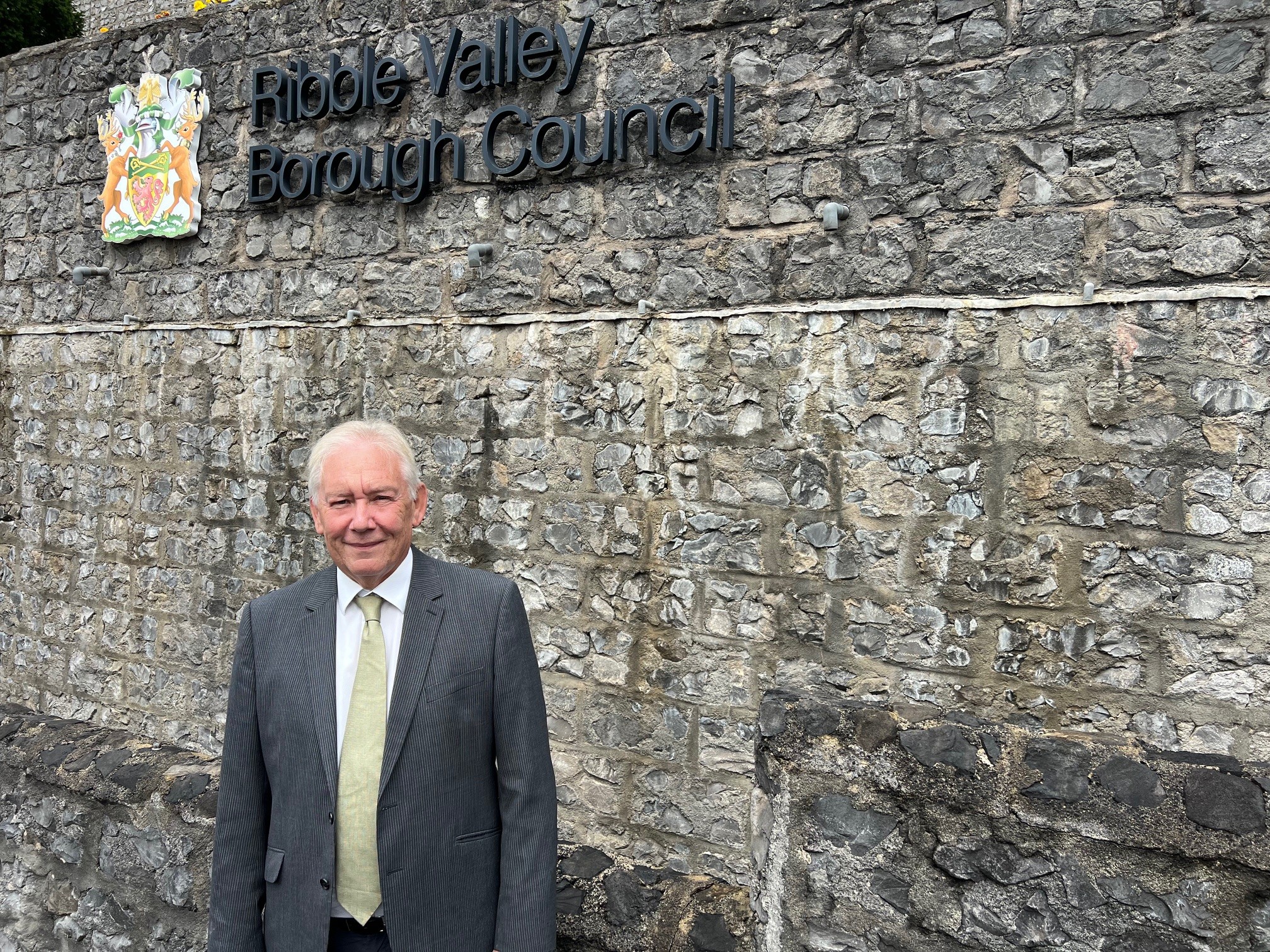 Retiring Ribble Valley Borough Council Chief Executive Marshal Scott stood in front of a grey stone wall with the RVBC logo